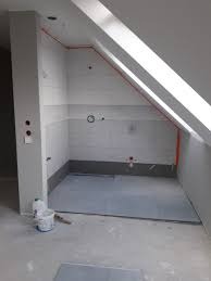 Surowa łazienka na poddaszu w trakcie remontu: częściowo wyłożone jasnoszare płytki na ścianach, przygotowane instalacje elektryczne i hydrauliczne, wylewka na podłodze, widoczny skośny sufit...