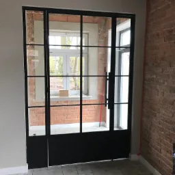 Metalowa konstrukcja z szybami w stylu loftowym, tworząca ściankę działową lub drzwi, widoczna na tle surowej, ceglanej ściany i okna z białymi ramami.