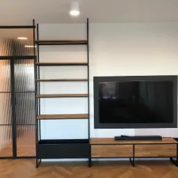 Nowoczesne wnętrze z elementami metalowymi: przeszklone drzwi z ryflowanego szkła w czarnej ramie, regał z metalową konstrukcją i drewnianymi półkami, szafka RTV z drewnianymi frontami i metalowymi...