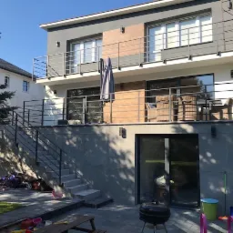 Dwukondygnacyjny dom z balkonami zabezpieczonymi metalowymi barierkami, schody zewnętrzne z metalową poręczą prowadzące na podwyższony taras, widok z ogrodu.