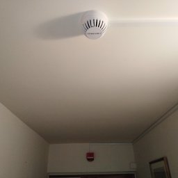 ELEKTRONET MARCIN BEREŚNIEWICZ - Zamontowany na suficie biały czujnik dymu z widocznymi szczelinami wentylacyjnymi i napisem 'CZUJNIK DYMU-2' w korytarzu budynku mieszkalnego, z widocznym fragmentem ściany, listwy sufitowej...