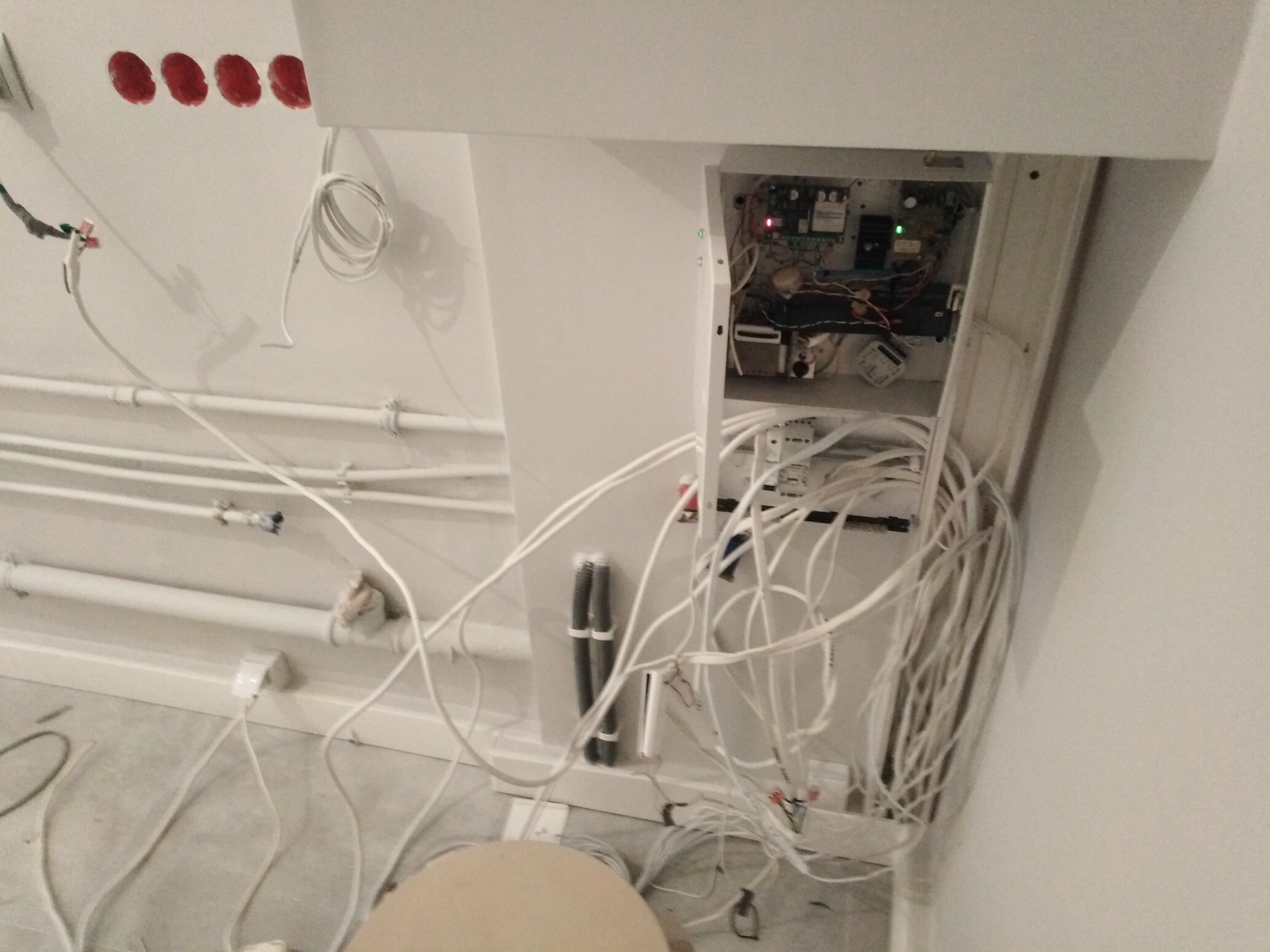 Otwarta skrzynka sterująca systemem detekcji alarmów technicznych z widocznymi przewodami i elektroniką, w tle rury instalacyjne na ścianie.