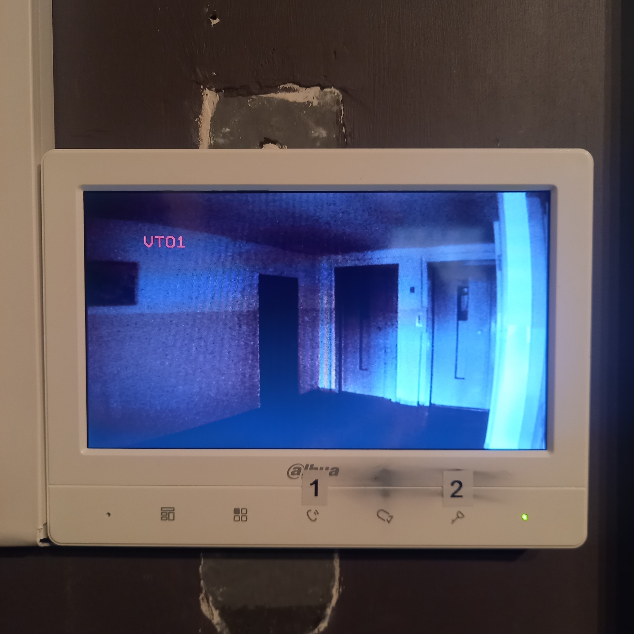Ekran videodomofonu marki Dahua, wyświetlający obraz z kamery zamontowanej wewnątrz budynku, widoczny korytarz z drzwiami i windą. Na obudowie naklejki z cyframi 1 i 2.