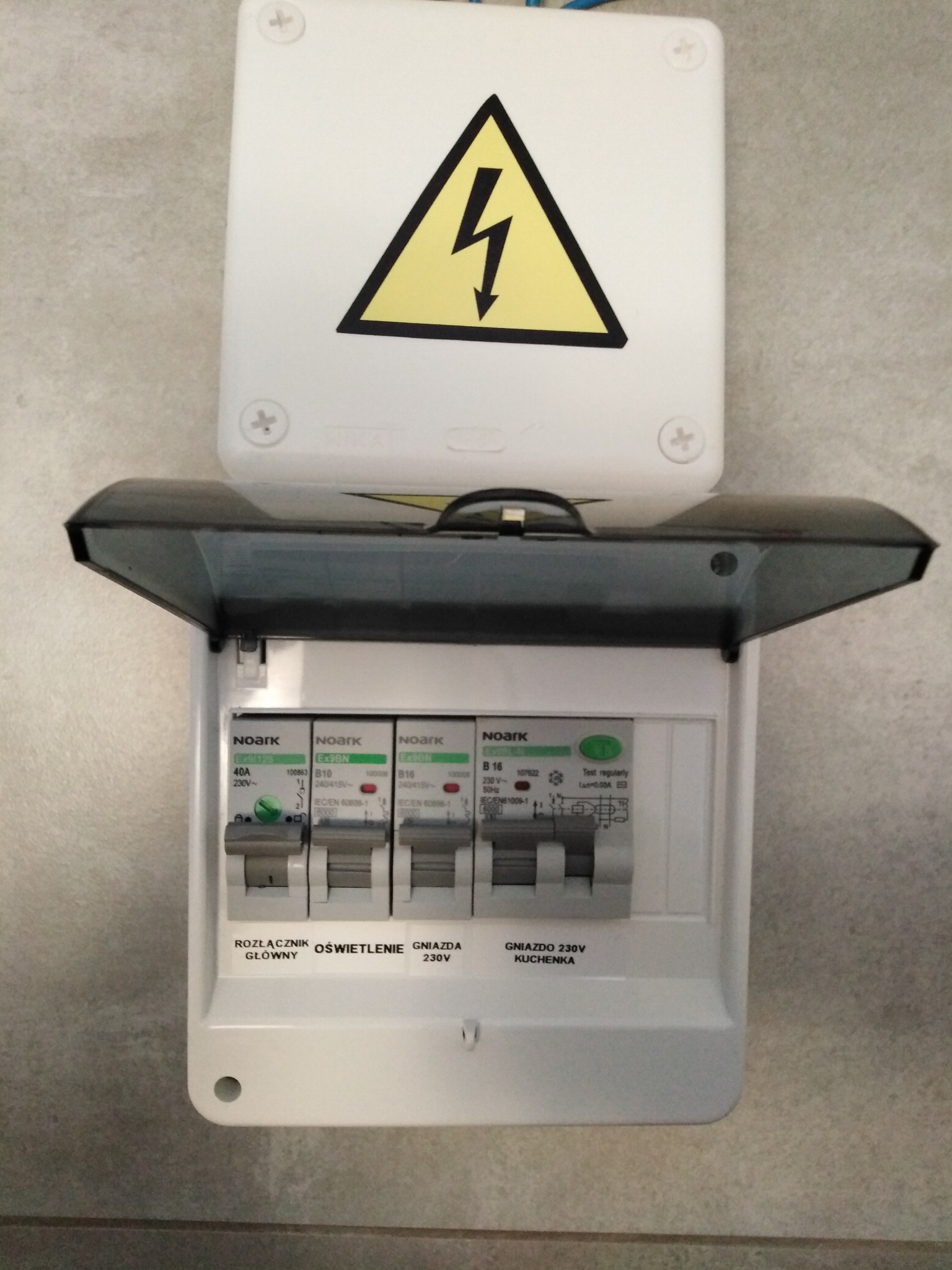 Otwarta skrzynka bezpiecznikowa Noark z widocznymi bezpiecznikami oznaczonymi jako rozłącznik główny, oświetlenie i gniazda 230V, w tym gniazdo kuchenki, z żółtym znakiem ostrzegawczym o wysokim...
