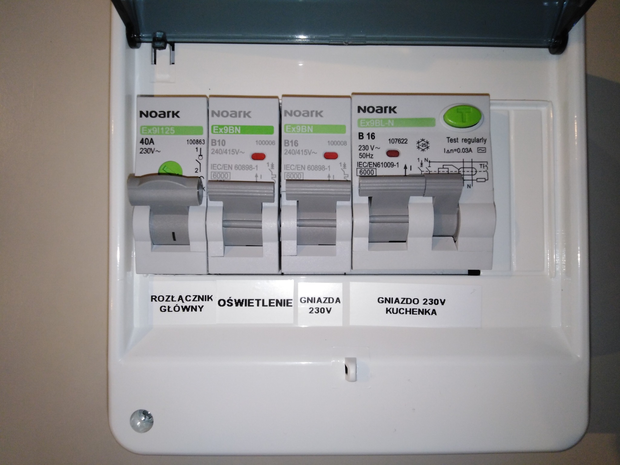 Otwarta skrzynka bezpiecznikowa marki Noark z czterema wyłącznikami, opisana etykietami: 'Rozłącznik główny', 'Oświetlenie', 'Gniazda 230V', 'Gniazdo 230V Kuchenka'.