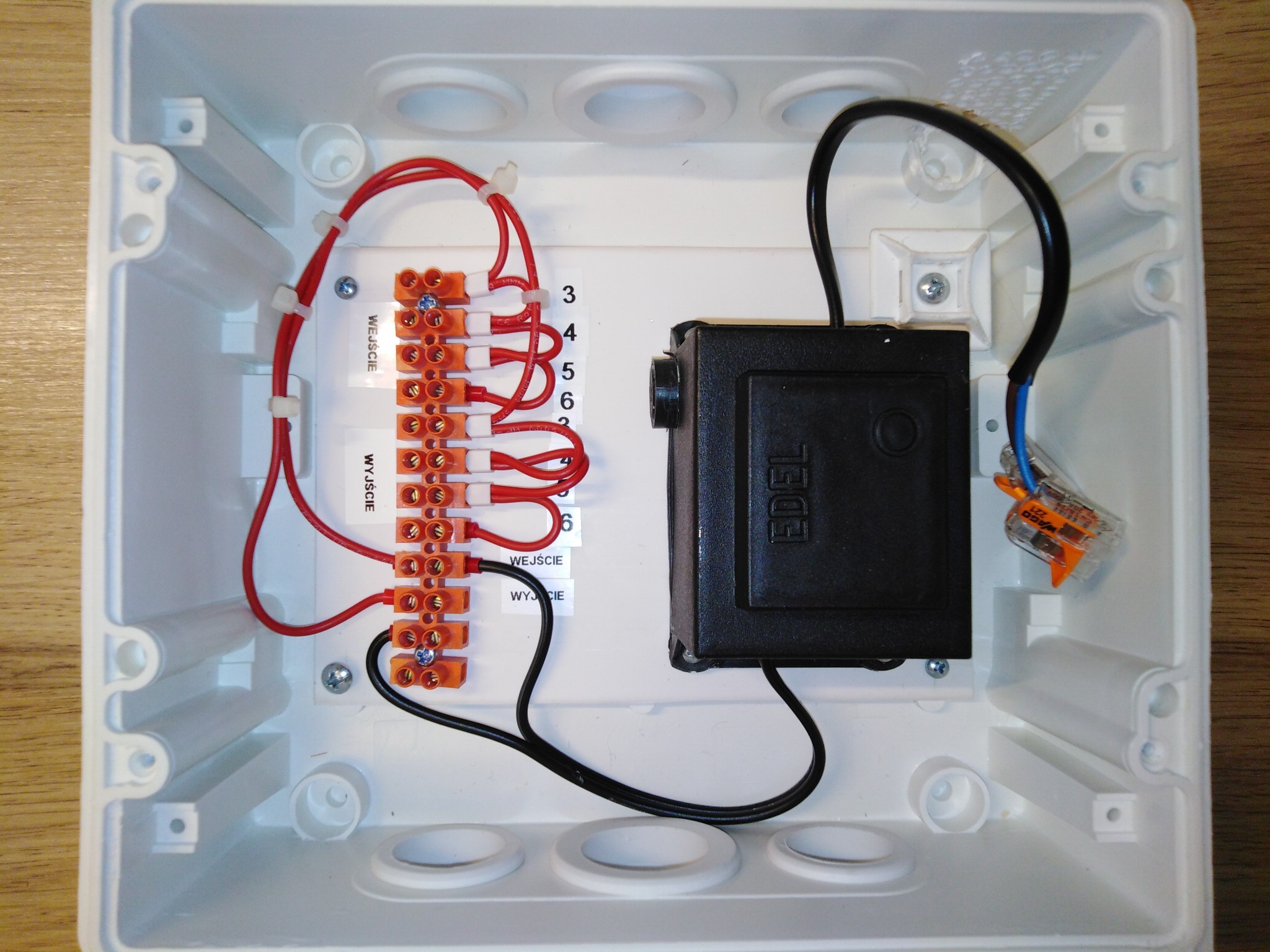 Wnętrze białej skrzynki elektrycznej z zamontowanymi listwami zaciskowymi, przewodami oznaczonymi jako 'Wejście' i 'Wyjście', czarnym transformatorem marki EDEL oraz złączką WAGO, całość...