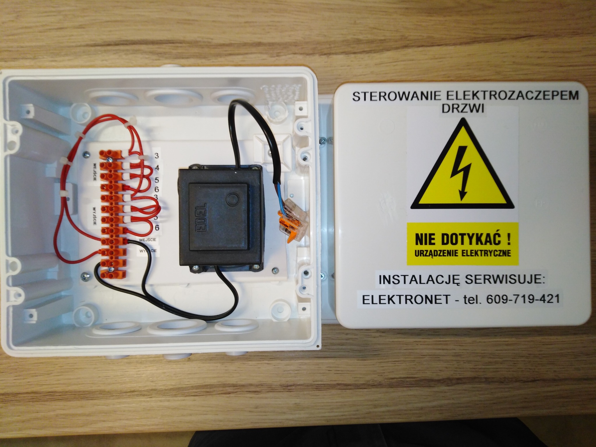 Otwarta skrzynka elektryczna sterowania elektrozaczepem drzwi z widocznymi przewodami, listwą zaciskową, transformatorem oraz naklejką ostrzegawczą 'Nie dotykać!' i danymi firmy serwisującej.