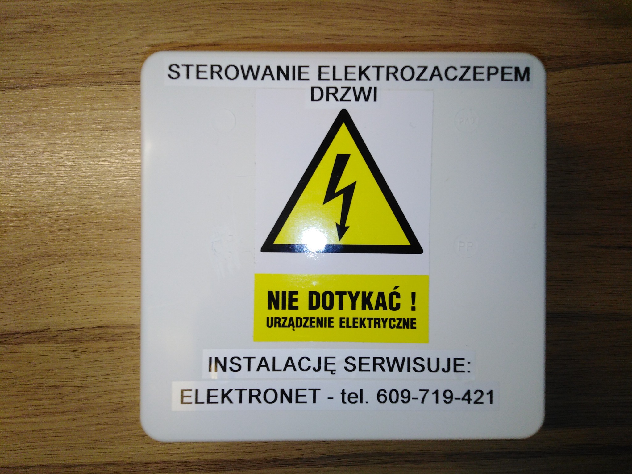 Biała skrzynka sterowania elektrozaczepem drzwi z ostrzeżeniem o napięciu, umieszczona na drewnianej powierzchni. Widoczna informacja o serwisowaniu instalacji przez firmę ELEKTRONET z numerem...