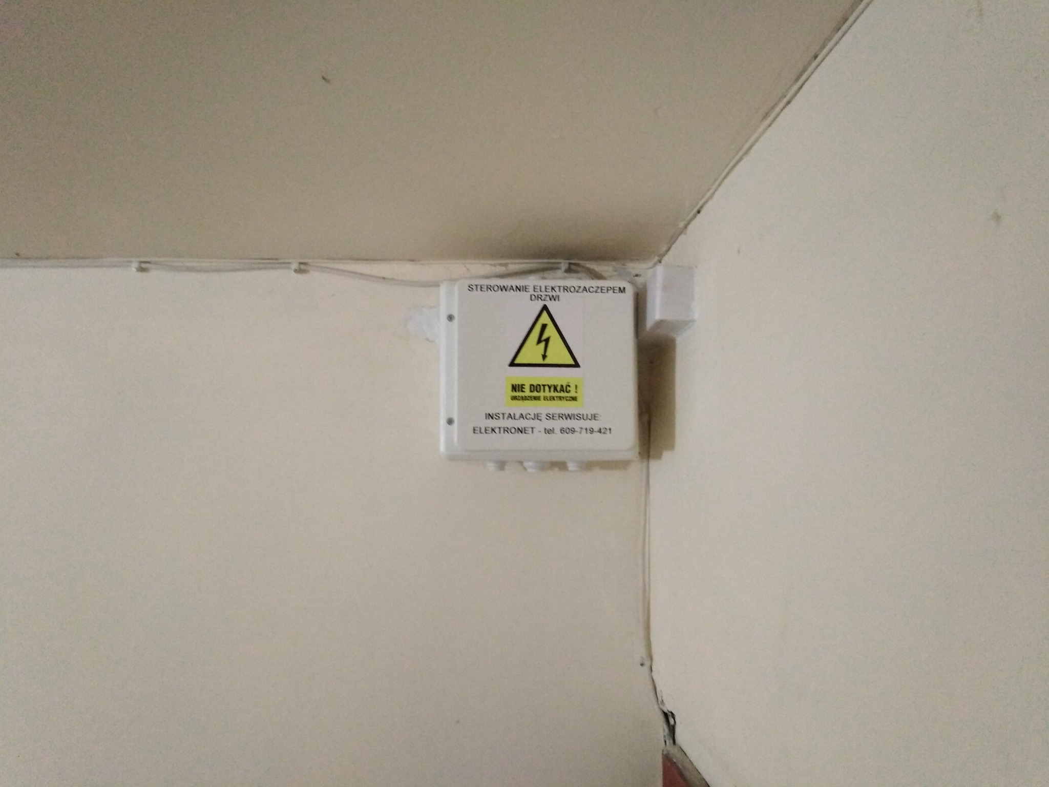 Zamknięta skrzynka elektryczna z napisem 'Sterowanie elektrozaczepem drzwi' i symbolem ostrzegawczym wysokiego napięcia, zamontowana na ścianie korytarza bloku mieszkalnego, z widocznymi przewodami...