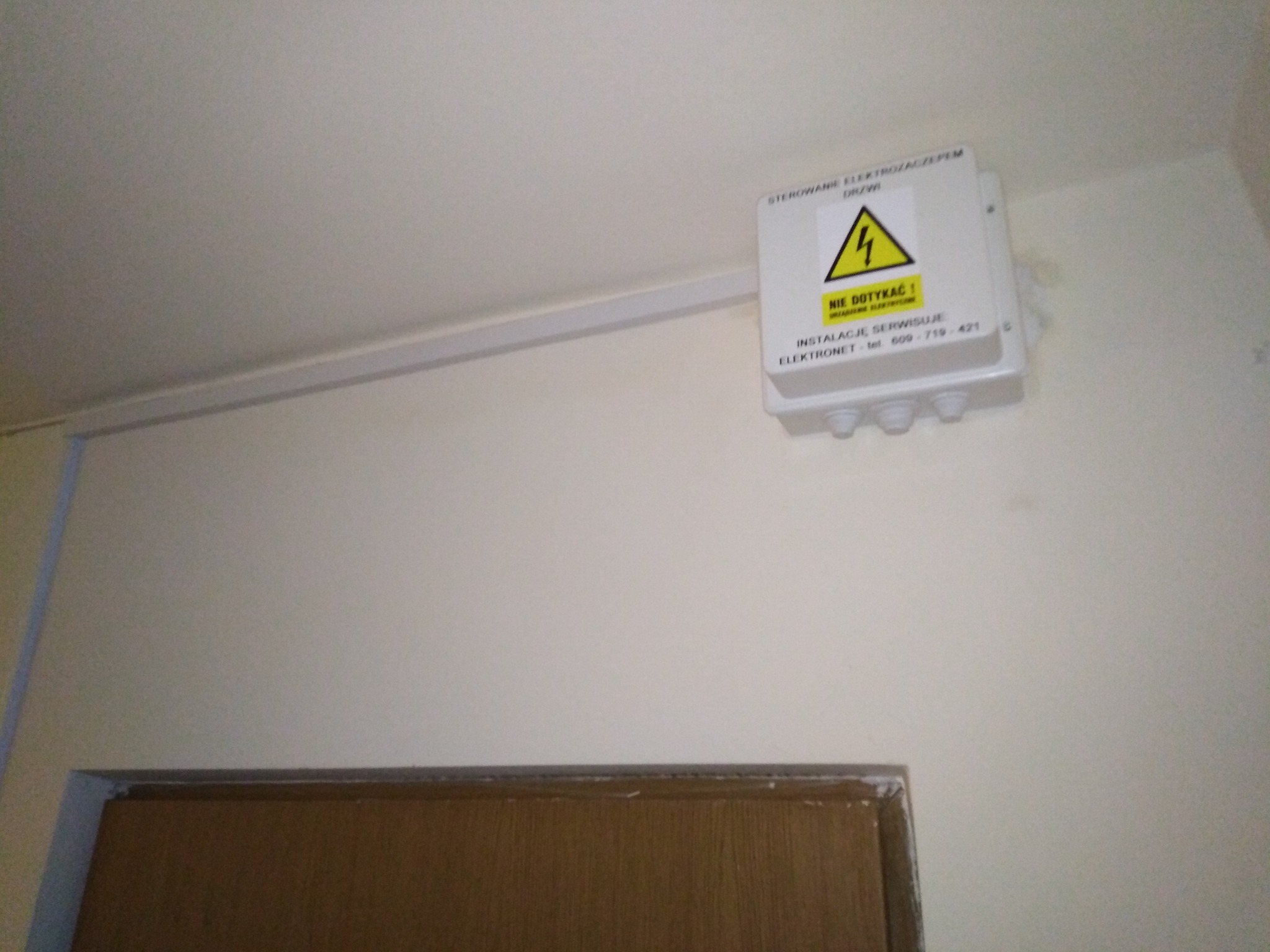 Biała puszka instalacyjna z napisem 'STEROWANE ELEKTROZACZEPEM DRZWI' i żółtym znakiem ostrzegawczym 'NIE DOTYKAĆ!', zamontowana na ścianie nad futryną drzwiową, z widoczną listwą kablową.