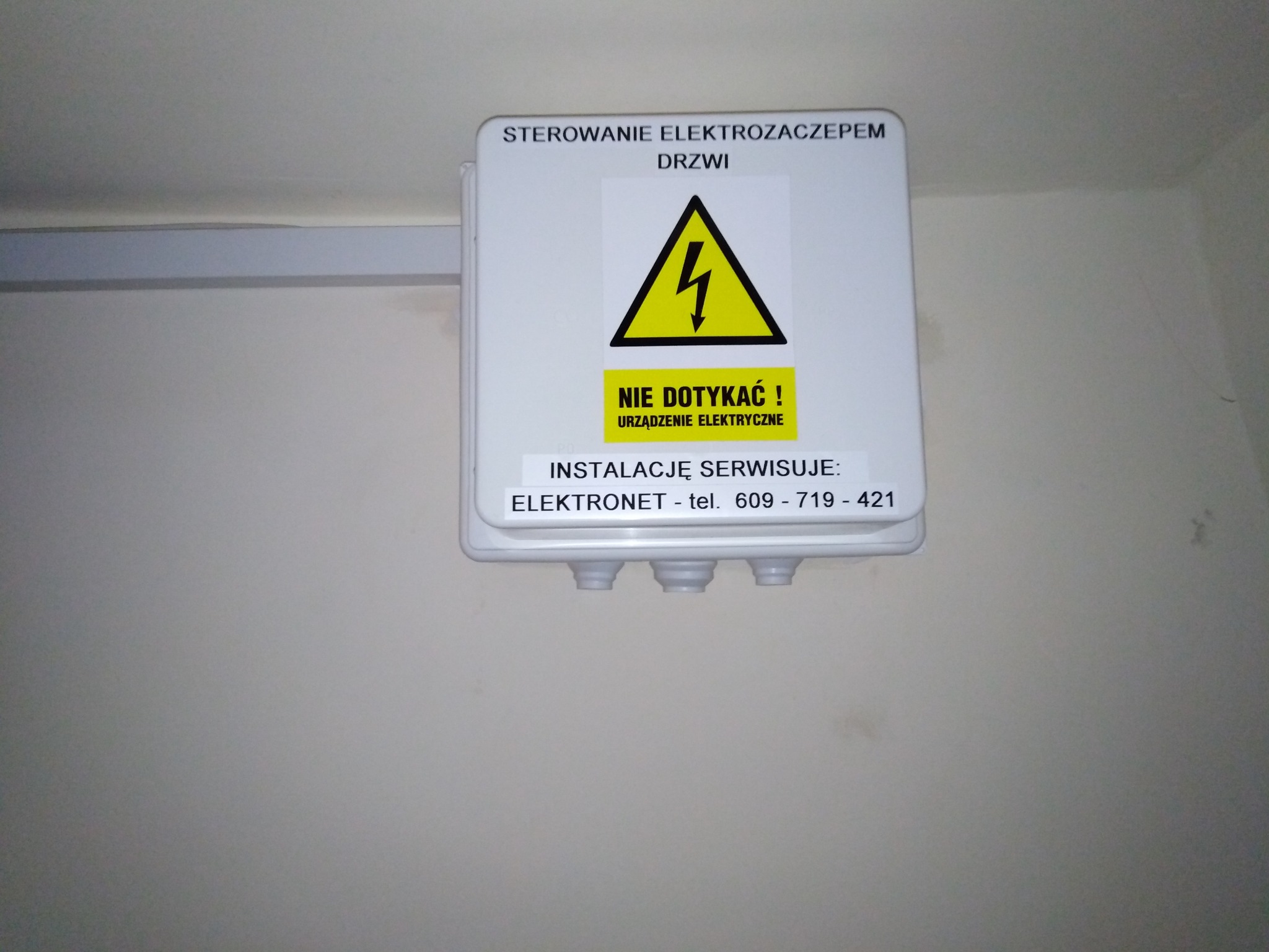 Biała skrzynka sterowania elektrozaczepem drzwi z żółtym znakiem ostrzegawczym o napięciu, przymocowana do ściany. Na skrzynce naklejka z numerem telefonu firmy serwisującej.