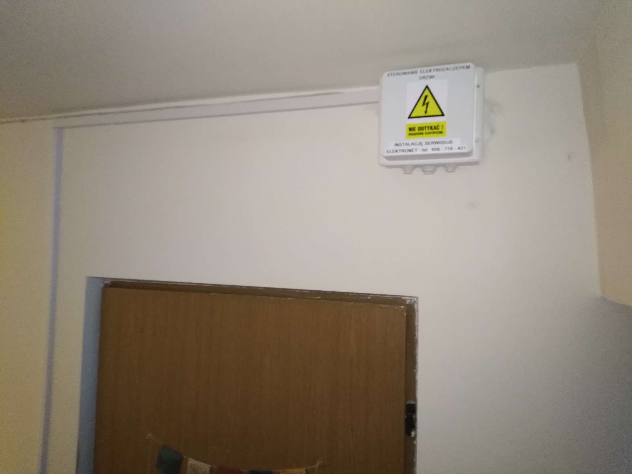 Biała skrzynka sterująca elektrozaczepem drzwi z żółtym znakiem ostrzegawczym wysokiego napięcia, zamontowana na białej ścianie nad brązowymi drzwiami.