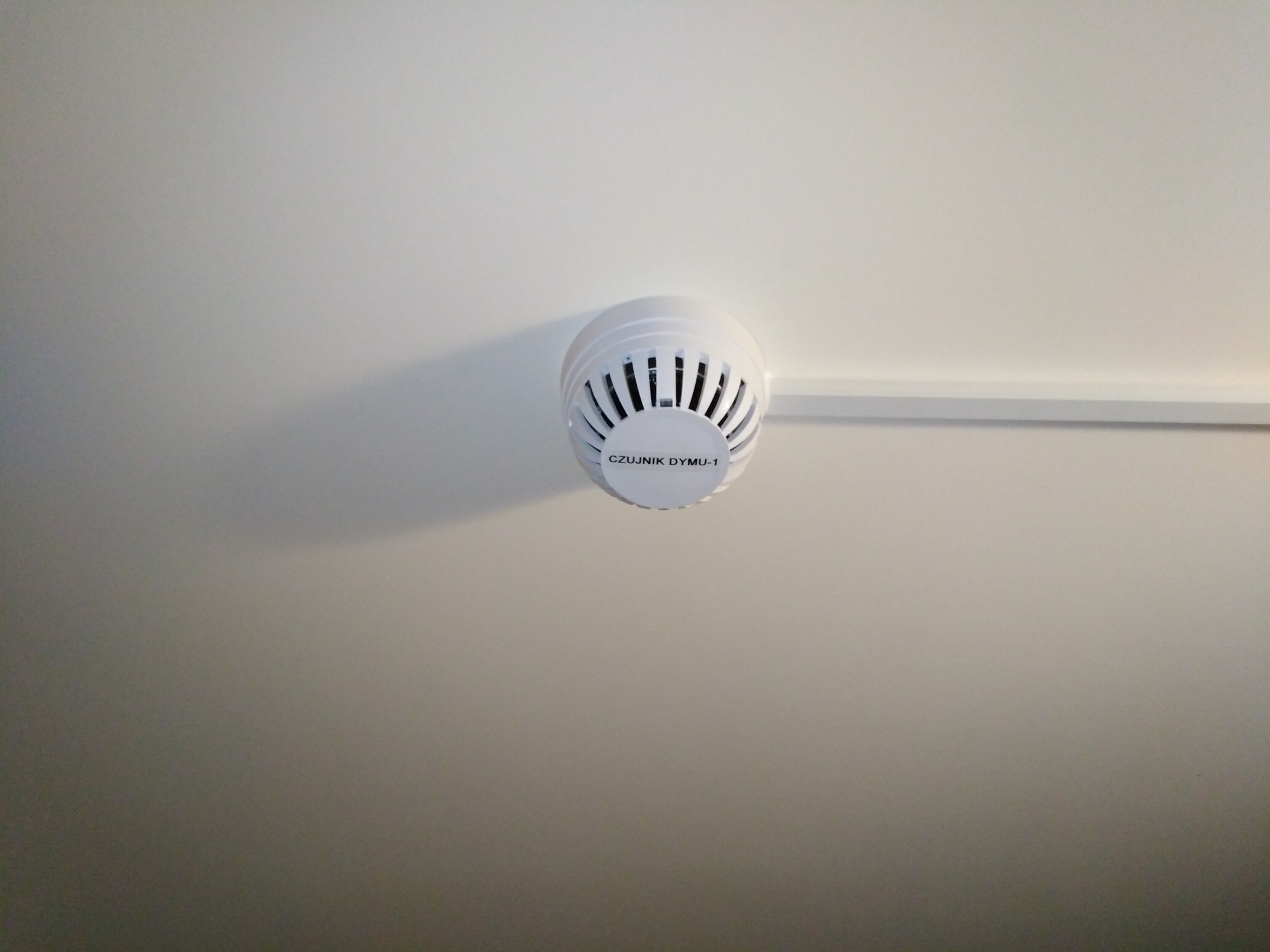 Biały czujnik dymu, model CZUJNIK DYMU-1, zamontowany na białym suficie, widoczna listwa przysufitowa.