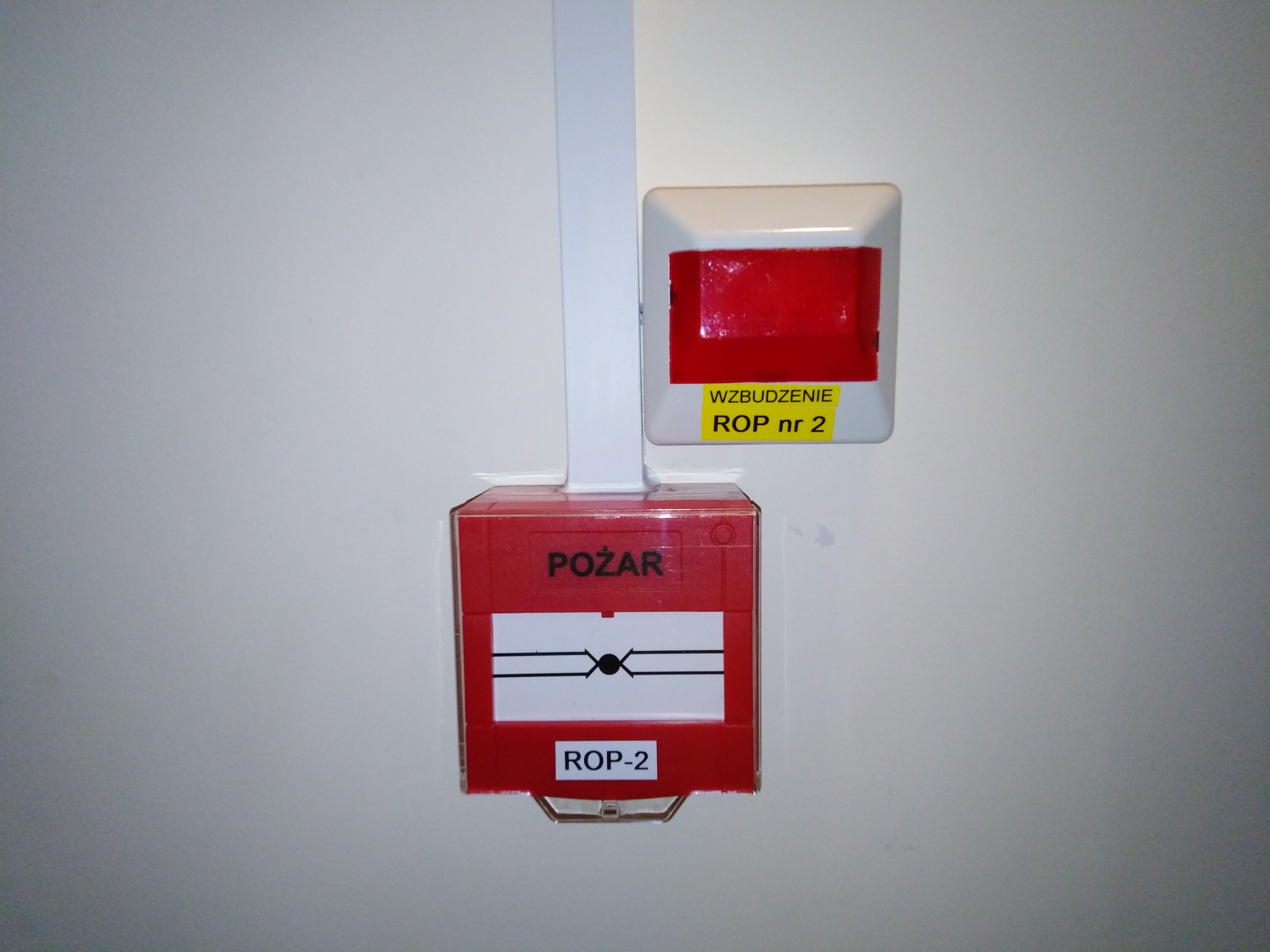 Czerwony ręczny ostrzegacz pożarowy ROP-2, zamontowany na białej ścianie korytarza, z żółtą tabliczką informującą o wzbudzeniu ROP nr 2, podłączony kablem w białej listwie.