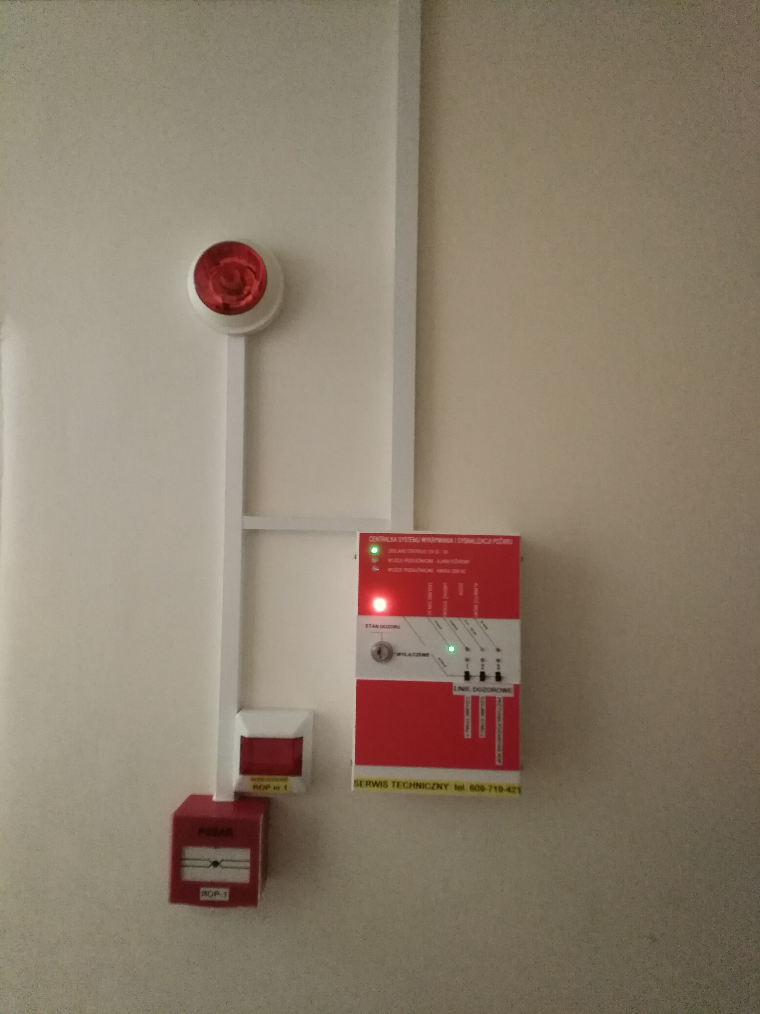 Zamontowany system sygnalizacji pożaru w korytarzu bloku mieszkalnego: centrala sterująca w czerwonej obudowie z diodami LED, czujnik dymu z sygnalizatorem akustycznym oraz ręczny ostrzegacz...