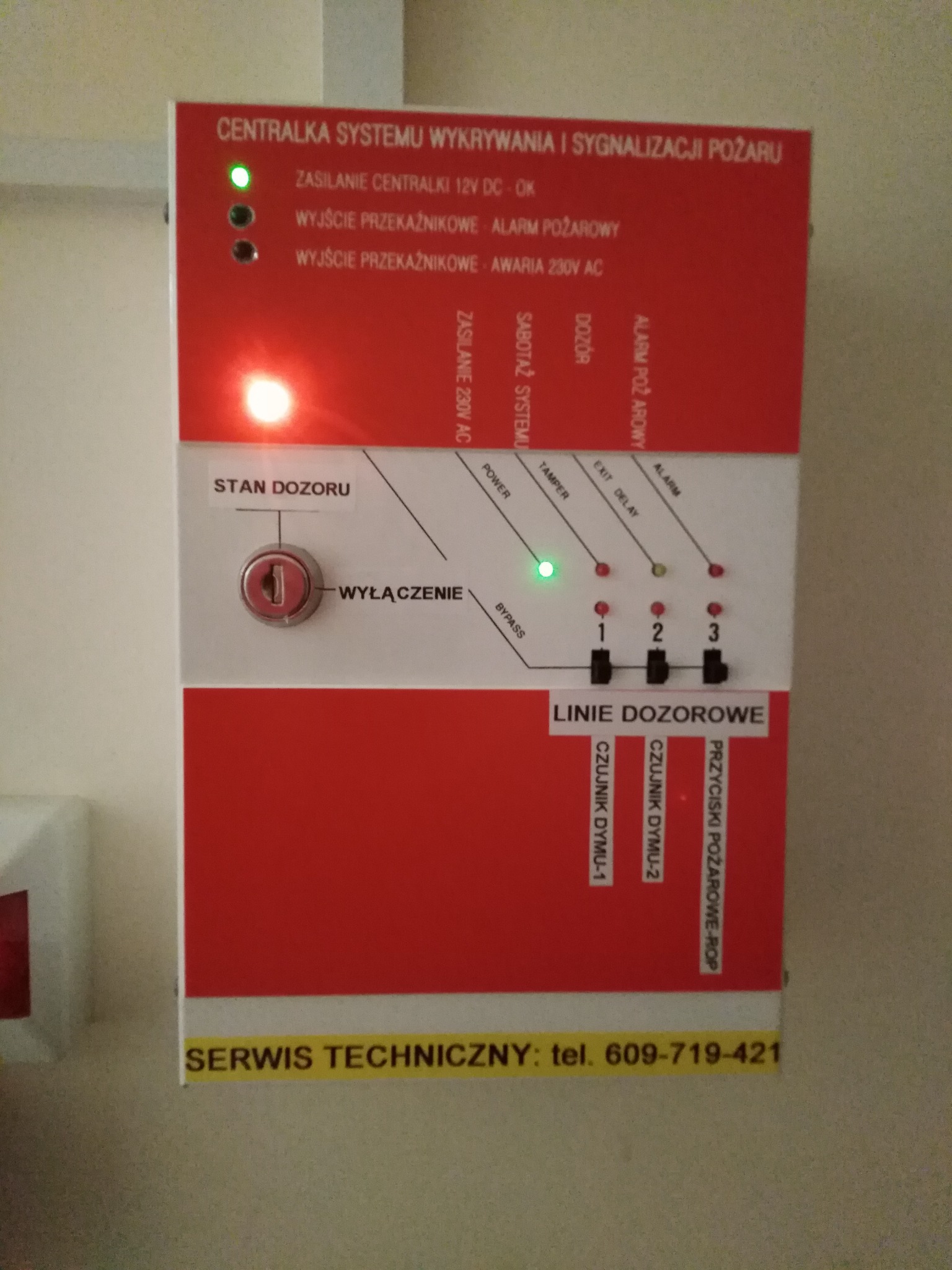 Centrala systemu wykrywania i sygnalizacji pożaru z włączonym zasilaniem, widoczne diody sygnalizujące stan dozoru, awarię i alarm, linie dozorowe podłączone do czujników dymu i przycisków ROP,...