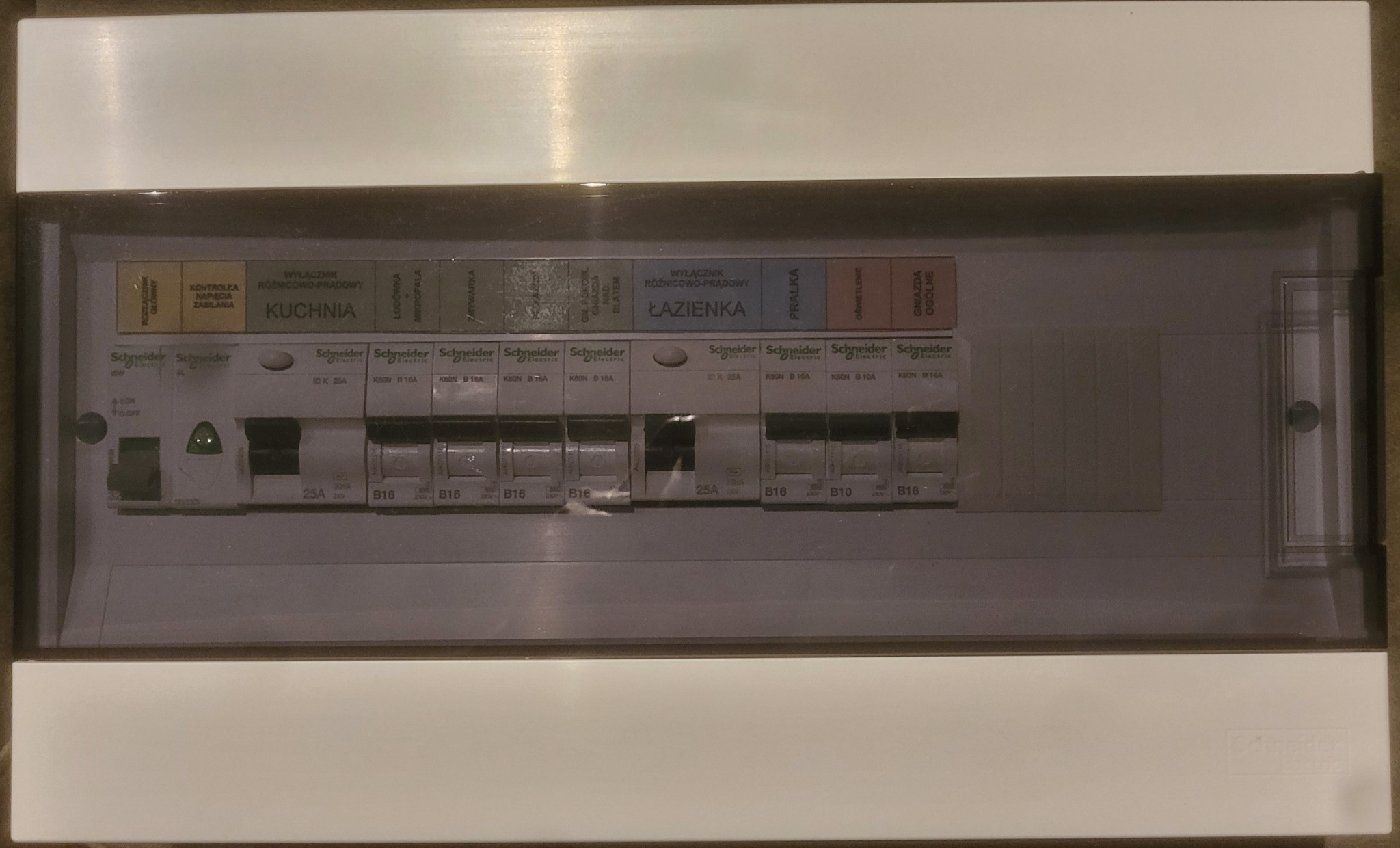 Rozdzielnica elektryczna Schneider z opisami obwodów (kuchnia, łazienka) i zabezpieczeniami B10/B16/25A, zamontowana w białej obudowie z przezroczystym panelem.