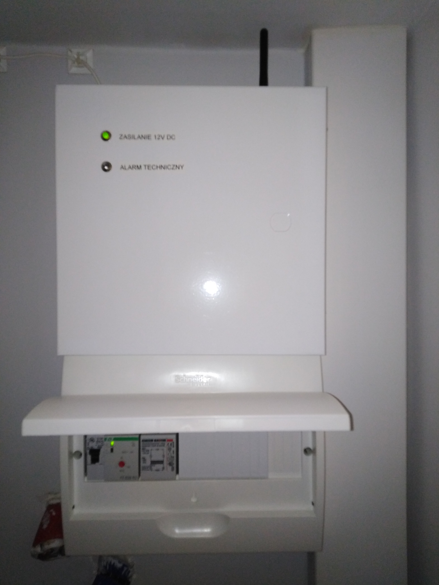 Biała centrala sterująca systemem detekcji alarmów technicznych z anteną, zamontowana na ścianie. Widoczny otwarty panel z elementami sterującymi i diodami.