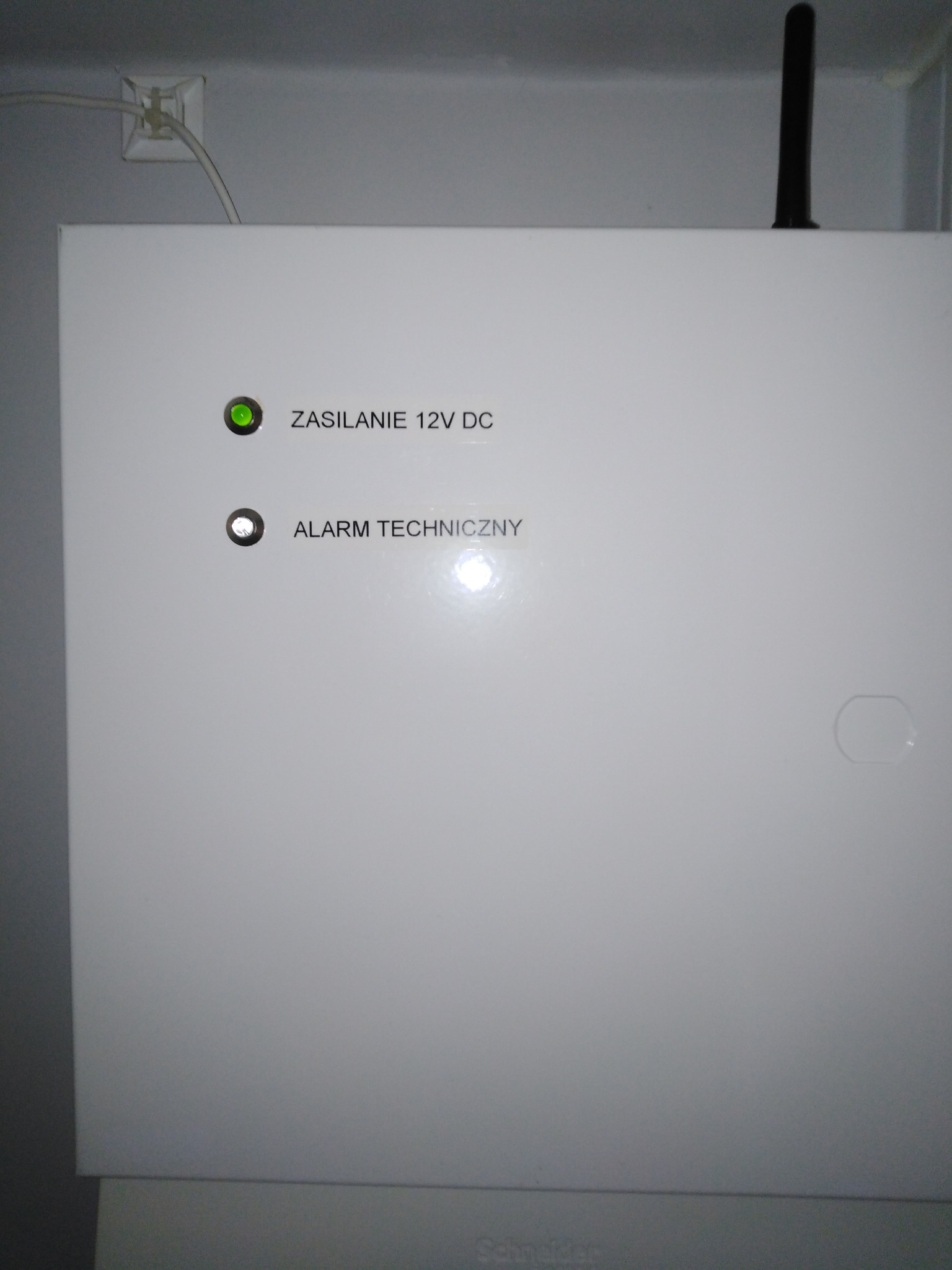Centrala alarmowa w białej obudowie z diodami sygnalizującymi zasilanie 12V DC i alarm techniczny, zamontowana na ścianie. Widoczna antena GSM.