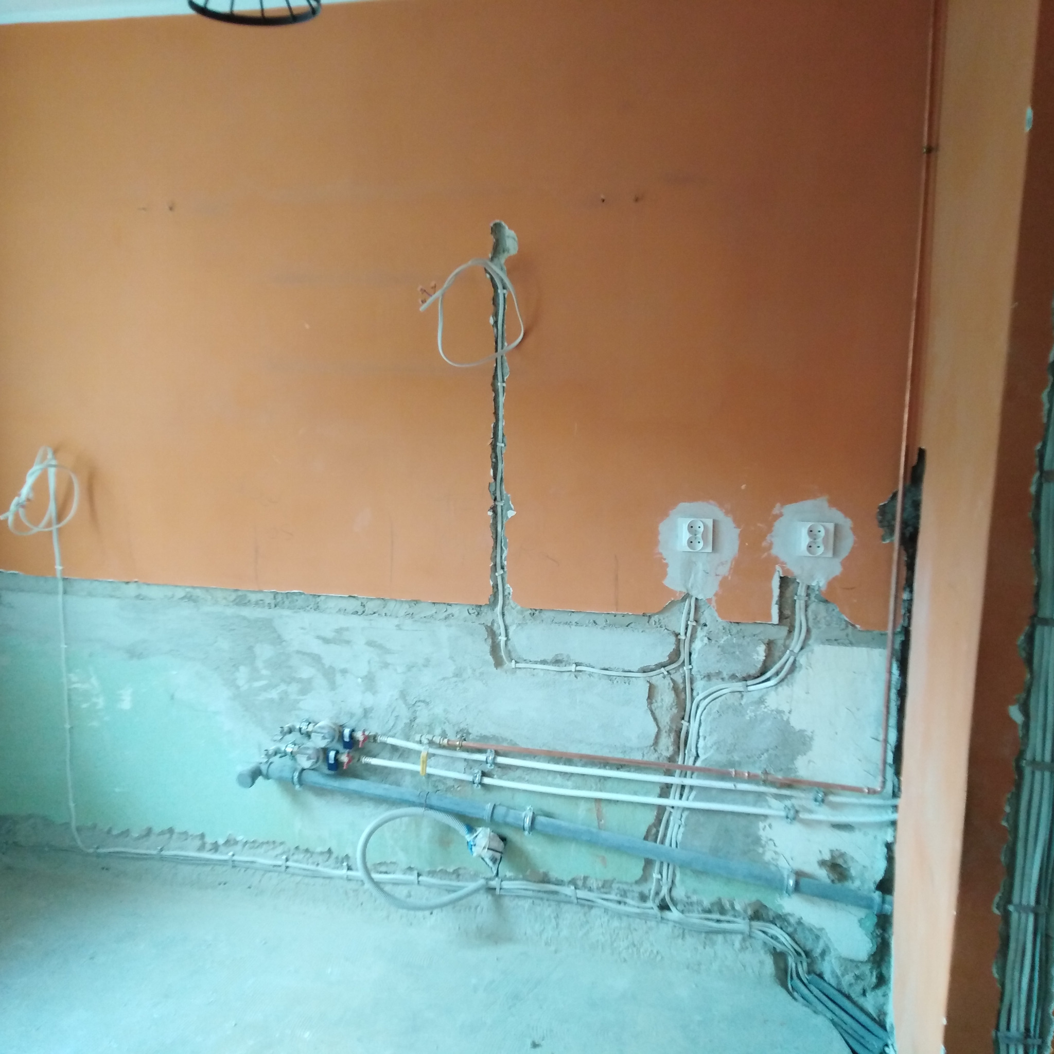 Pomarańczowa ściana w trakcie remontu z widocznymi instalacjami elektrycznymi i hydraulicznymi. Gniazdka elektryczne osadzone w ścianie, rury i kable prowadzone w bruzdach.