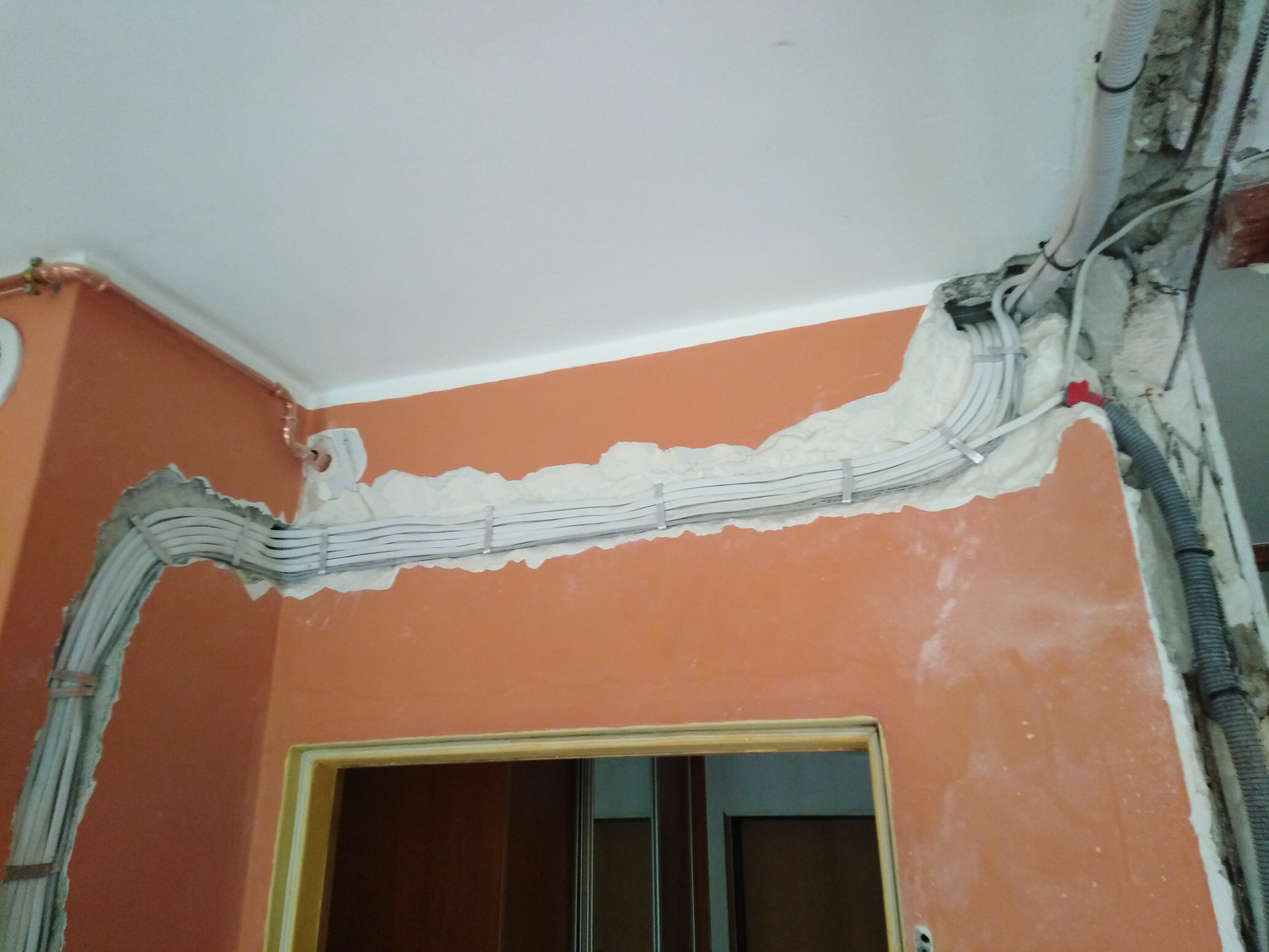 Bruzdy w ścianie z poprowadzonymi przewodami elektrycznymi, widoczne kable w białych rurkach, fragment sufitu i ościeżnica drzwiowa. Pomarańczowa ściana w trakcie prac.