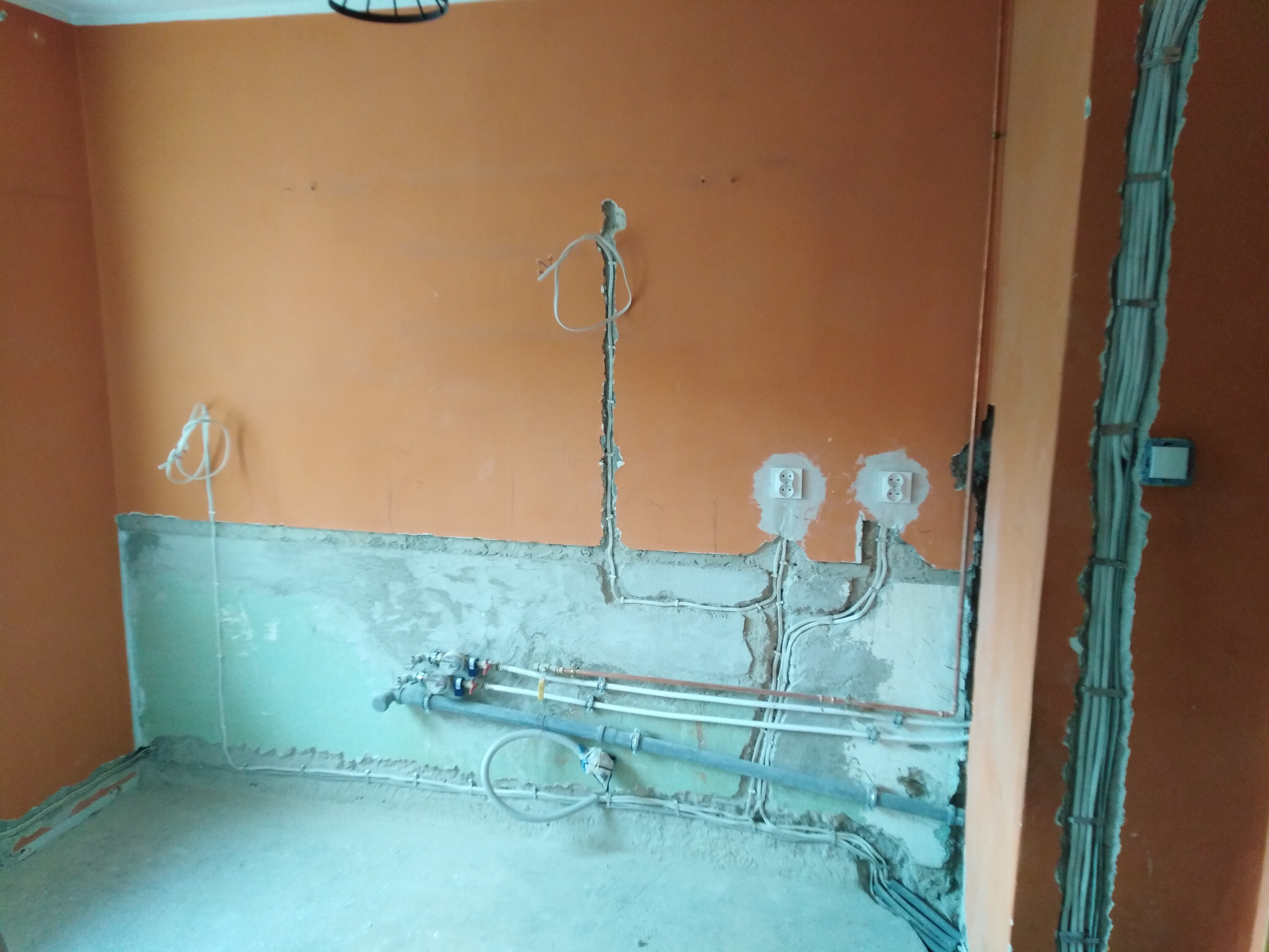 Pomarańczowa ściana z widocznymi przewodami elektrycznymi i rurami w trakcie instalacji. Widoczne puszki elektryczne i fragment instalacji w ścianie.