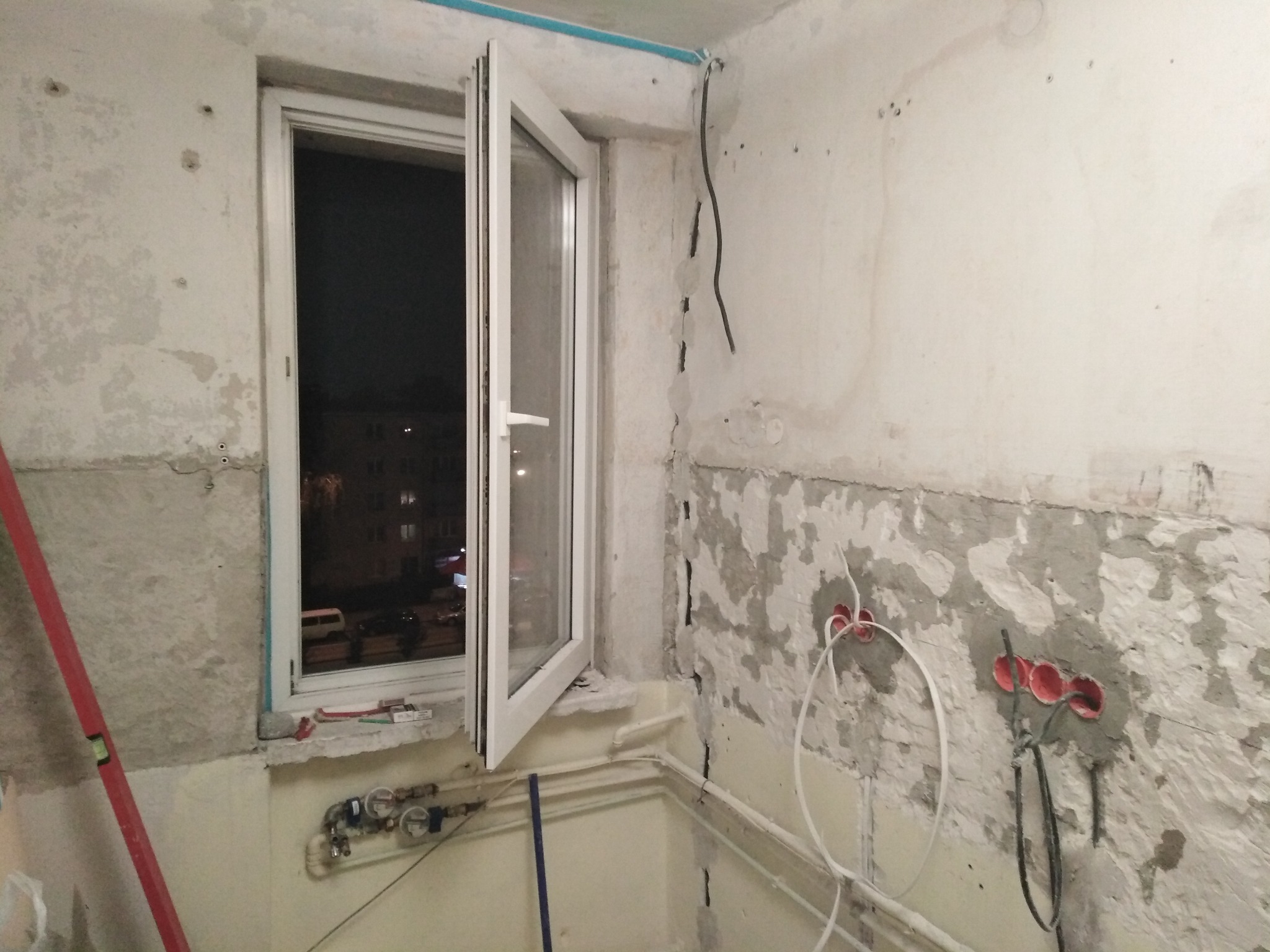 Wnętrze kuchni w trakcie modernizacji instalacji elektrycznej w Gdańsku. Widoczne kable wystające ze ściany z przygotowanymi otworami pod gniazdka, okno otwarte na nocny widok miasta oraz poziomica...