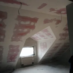 Poddasze w trakcie prac wykończeniowych: ściany i sufit pokryte warstwą szpachli, widoczne kable wystające z sufitu, okno w skosie z zaklejonym grzejnikiem, surowa podłoga.