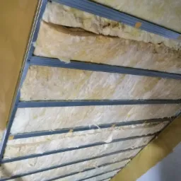 Skośny sufit poddasza z widoczną wełną mineralną pomiędzy metalowymi profilami, częściowo zabezpieczoną żółtą folią paroizolacyjną.