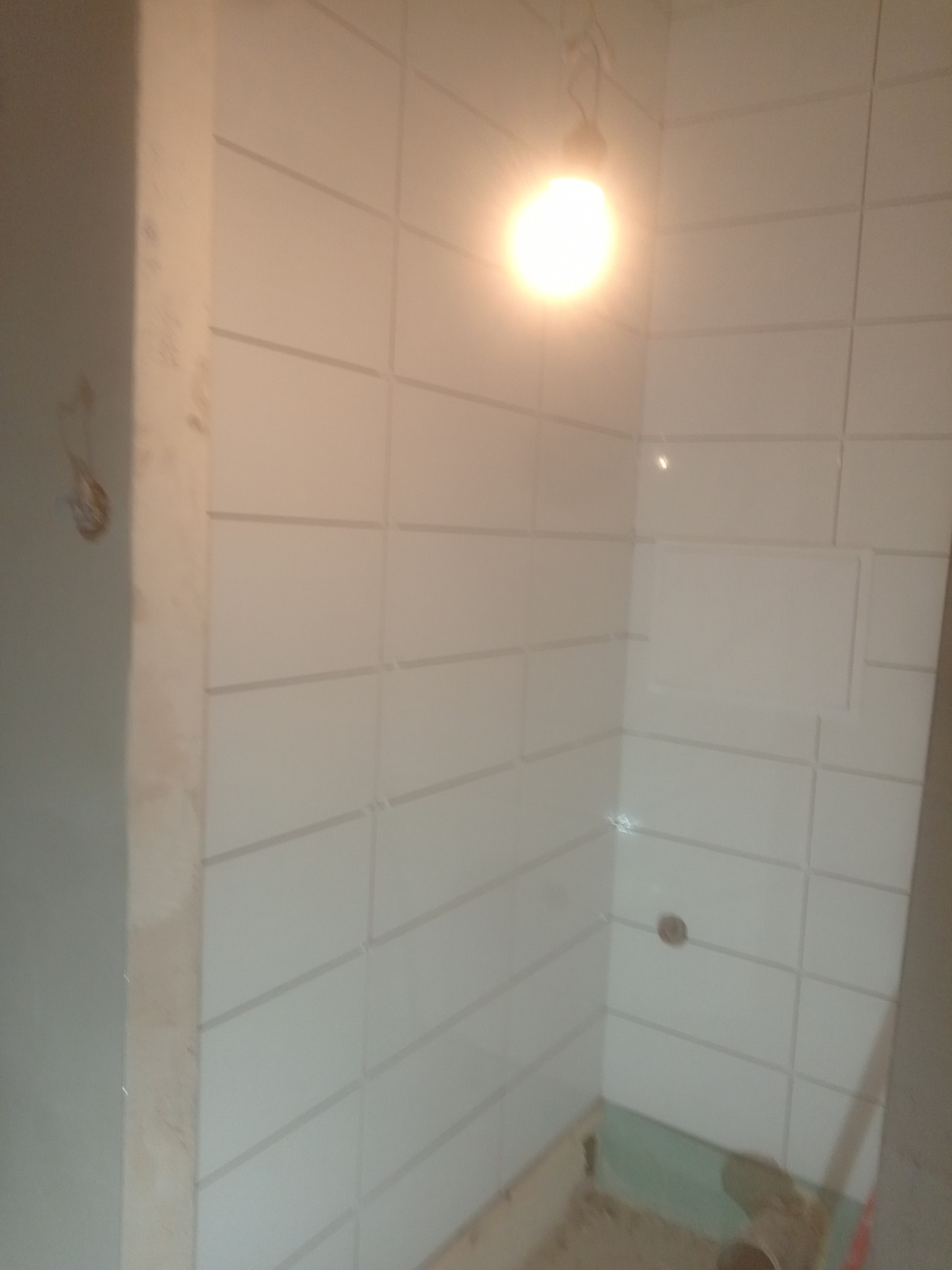 Świeżo wykafelkowana, biała ściana w łazience, oświetlona wiszącą żarówką, z widocznym otworem na rurę i fragmentem podłogi w trakcie prac wykończeniowych.