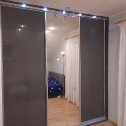 Nowoczesna szafa przesuwna z szarymi frontami i lustrzanym panelem, oświetlenie LED na górze, widok na sypialnię z łóżkiem w odbiciu lustra.