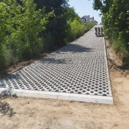 Długa, lekko wznosząca się droga z ażurowej kostki, obrzeża z jasnoszarego betonu, po bokach piasek i zieleń, w tle dom.