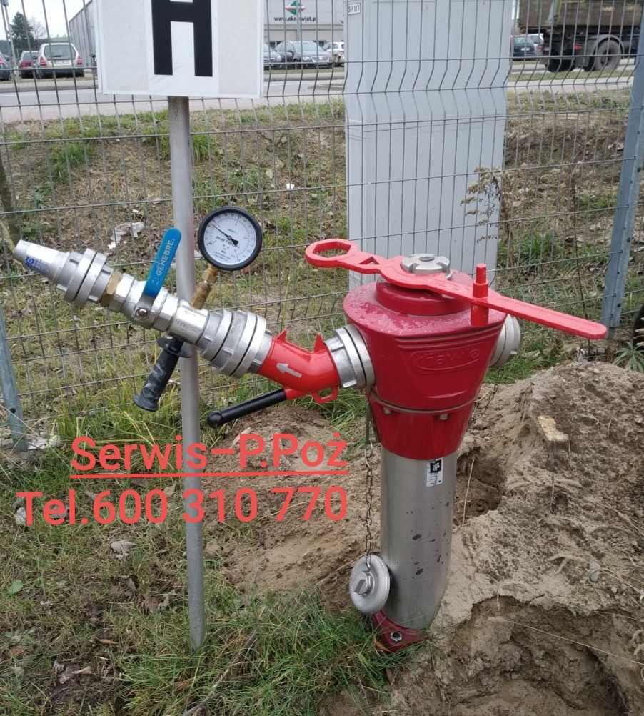 Czerwony hydrant naziemny z manometrem, z nasadą i kluczem, zainstalowany w ziemi, widoczny znak 'H' na słupku.