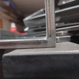 Stos profili stalowych kątowych, widoczne ślady obróbki mechanicznej na krawędziach, ułożone na metalowym blacie.