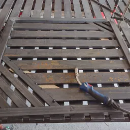 Metalowa konstrukcja bramy z poziomymi elementami i ukośnymi wzmocnieniami, częściowo zardzewiała, w trakcie spawania, widoczny uchwyt spawarki, zacisk stolarski i szczypce boczne.