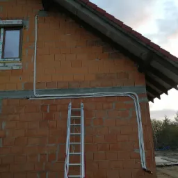 Surowe ściany z cegły domu jednorodzinnego z zewnętrznymi rurami wentylacyjnymi zamontowanymi na elewacji, widoczna drabina oparta o ścianę.