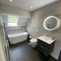 Nowoczesna łazienka na poddaszu z wolnostojącą wanną, umywalką z szafką w kolorze ciemnego drewna, podświetlanym lustrem i szarą glazurą na ścianach.