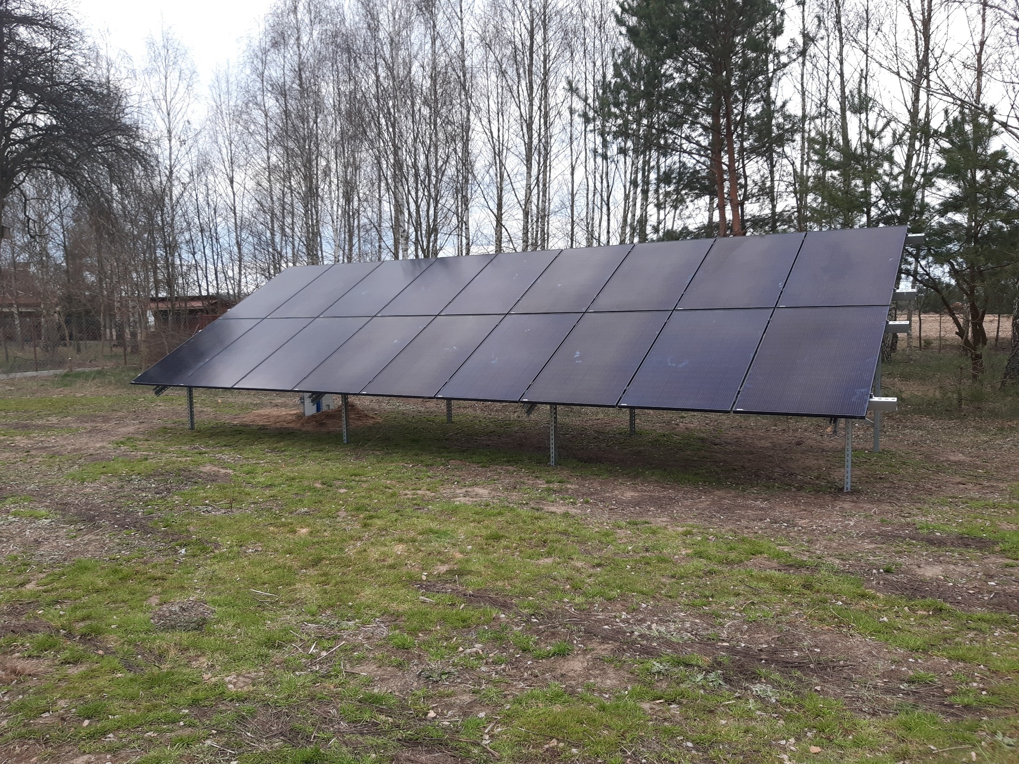 Rząd paneli słonecznych na metalowych wspornikach, ustawiony na trawiastym terenie w otoczeniu drzew.