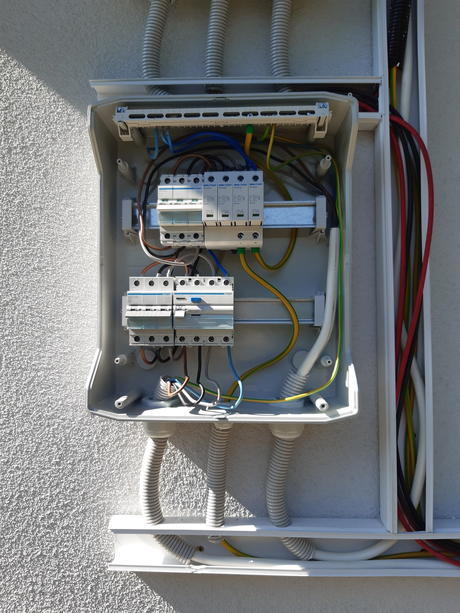 Otwarta skrzynka elektryczna z zainstalowanymi bezpiecznikami i przewodami, zamontowana na szorstkiej, pomalowanej ścianie zewnętrznej budynku, z widocznymi rurami karbowanymi doprowadzającymi kable.