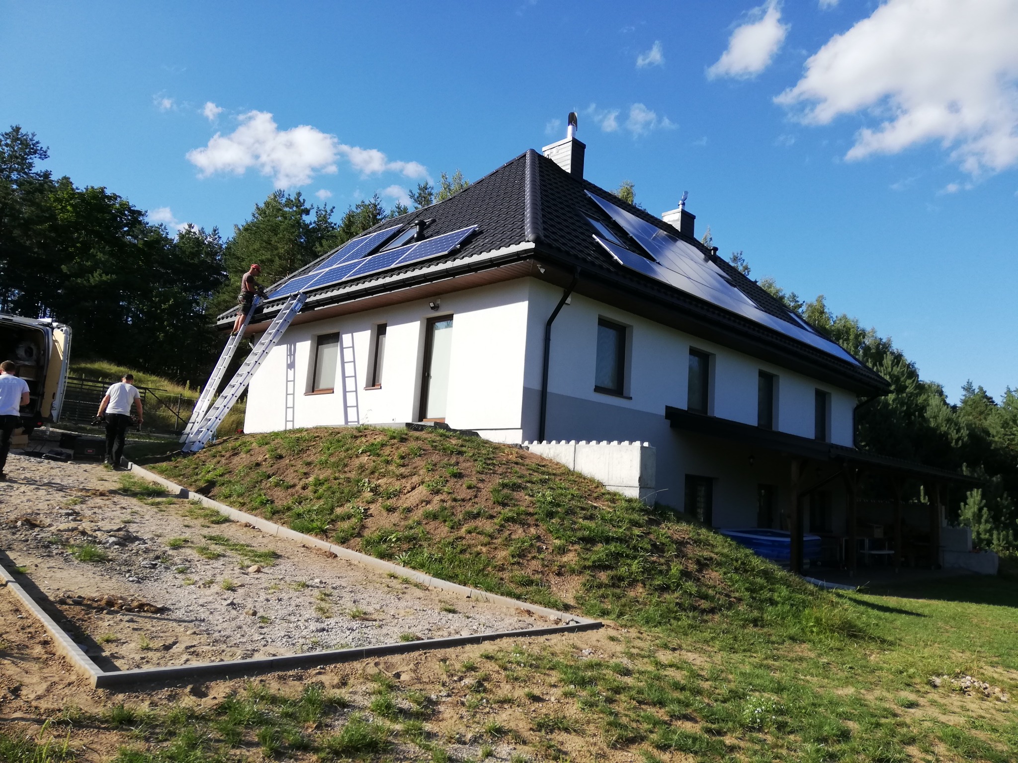 Montaż paneli słonecznych na dachu dwukondygnacyjnego domu jednorodzinnego z ciemną dachówką, widoczny pracownik na drabinie, częściowo zagospodarowany teren wokół budynku.