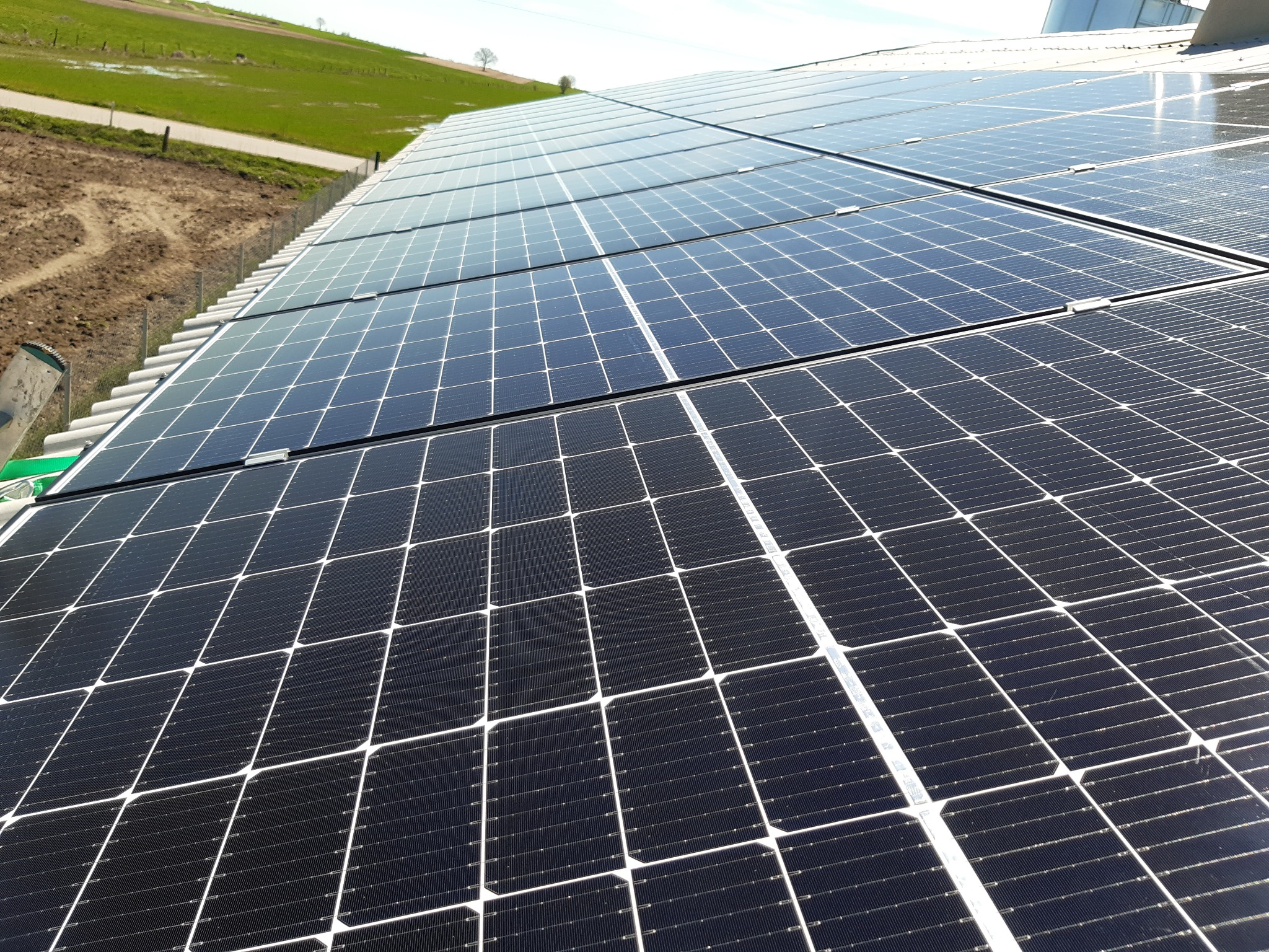 Rząd paneli fotowoltaicznych na dachu budynku, widok z dołu pod kątem, z zielonym polem i niebem w tle.