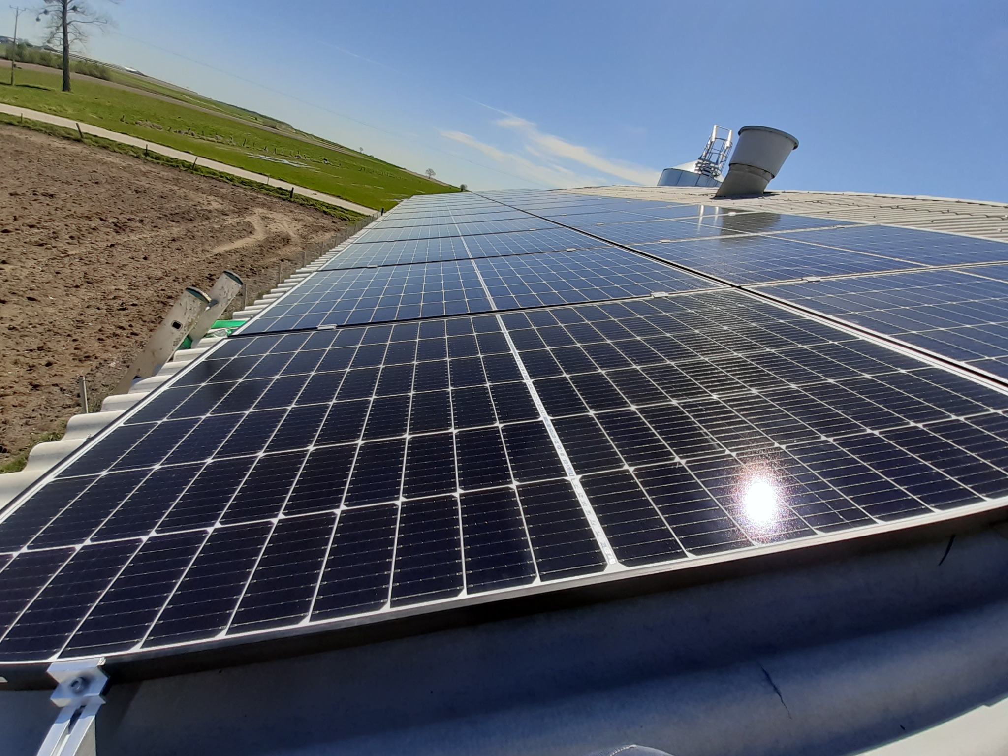 Rząd paneli słonecznych na metalowym dachu budynku gospodarczego, z drabinką i kominem wentylacyjnym w tle, na tle błękitnego nieba i zielonych pól.