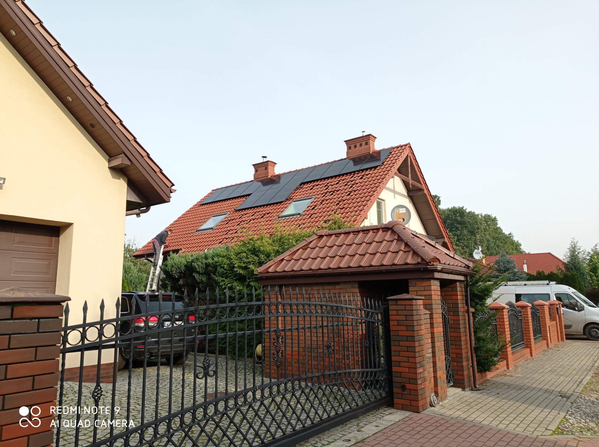 Dom jednorodzinny z czerwonym dachem, na którym zamontowano czarne panele słoneczne; mężczyzna na drabinie przy dachu, prawdopodobnie podczas montażu lub serwisu paneli.