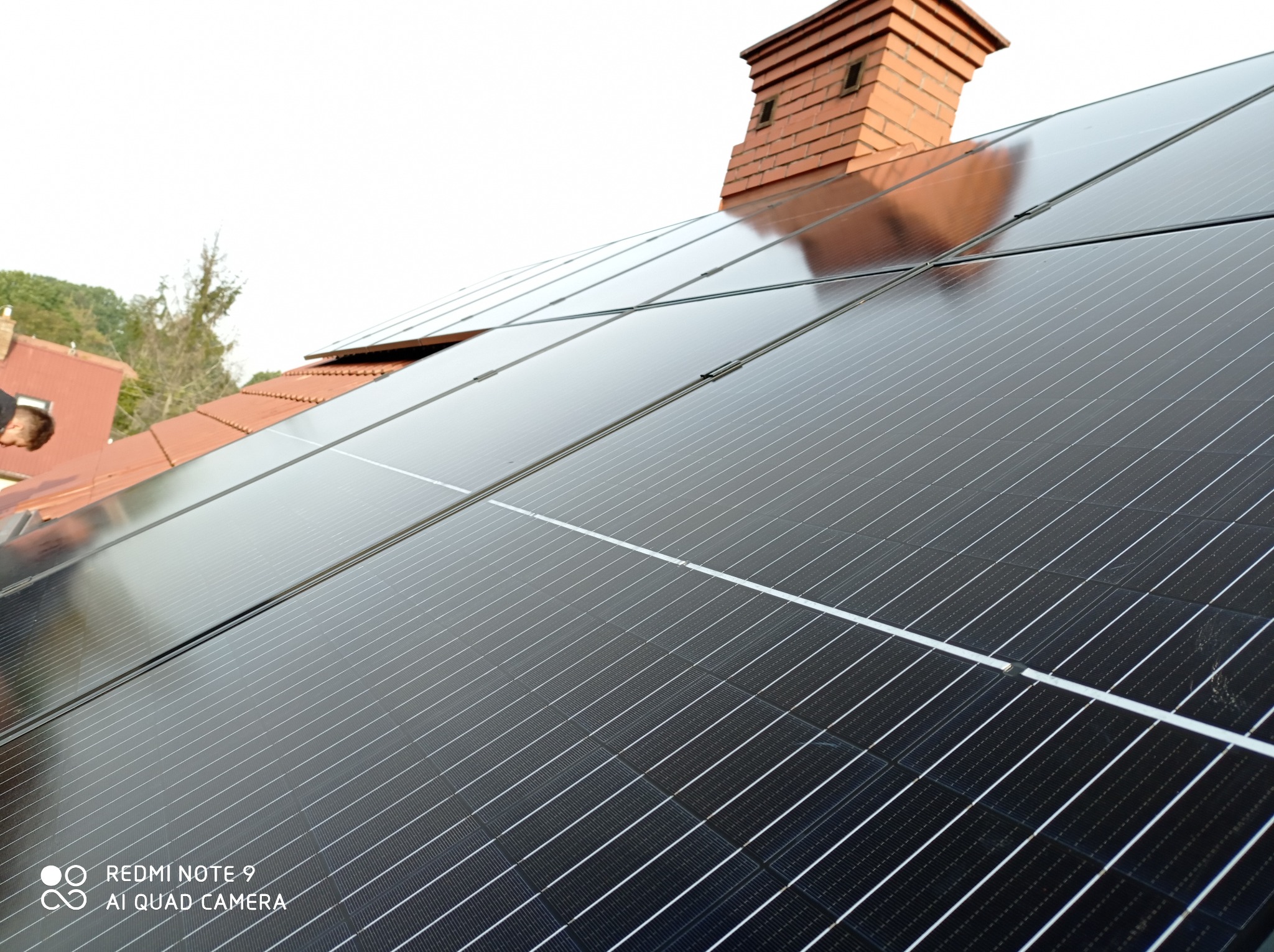 Częściowy widok instalacji paneli słonecznych na dachu z ceglanym kominem w tle, uchwycony pod kątem z widocznym fragmentem osoby po lewej stronie.