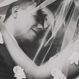 Czarno-białe zbliżenie pary młodej, delikatnie przesłoniętej koronkowym welonem, z subtelnym uśmiechem na twarzach i złączonymi czołami.
