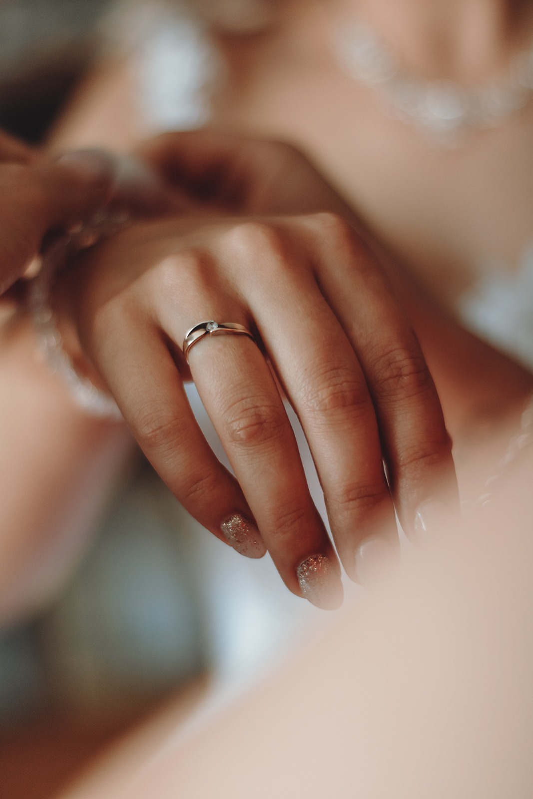 Dłoń kobiety z pierścionkiem zaręczynowym z pojedynczym diamentem i brokatowym manicure, delikatne koronki w tle.