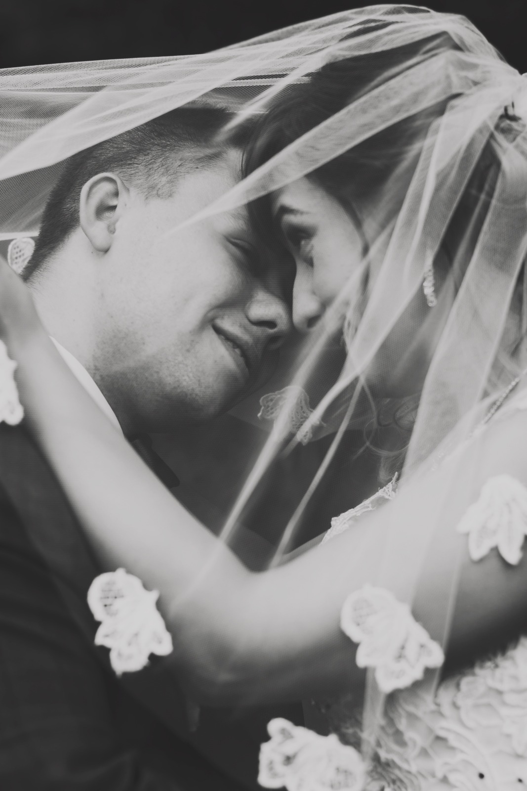 Czarno-białe zbliżenie pary młodej, delikatnie przesłoniętej koronkowym welonem, z subtelnym uśmiechem na twarzach i złączonymi czołami.