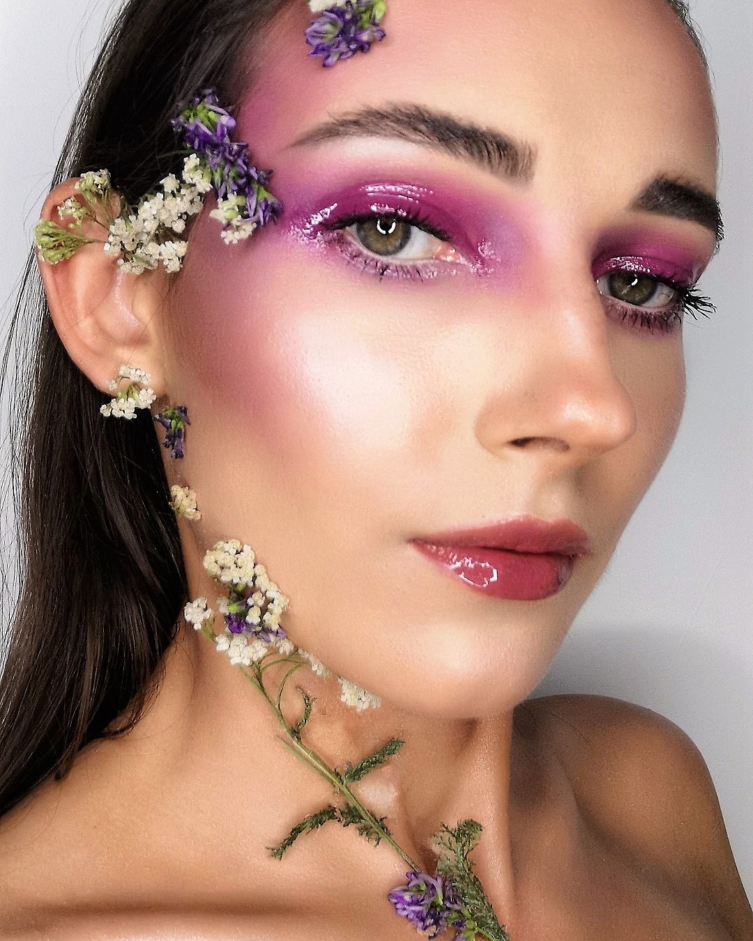 Portret kobiety z intensywnym fioletowym cieniem do powiek, błyszczącymi ustami i dekoracją z drobnych kwiatów na twarzy i szyi.