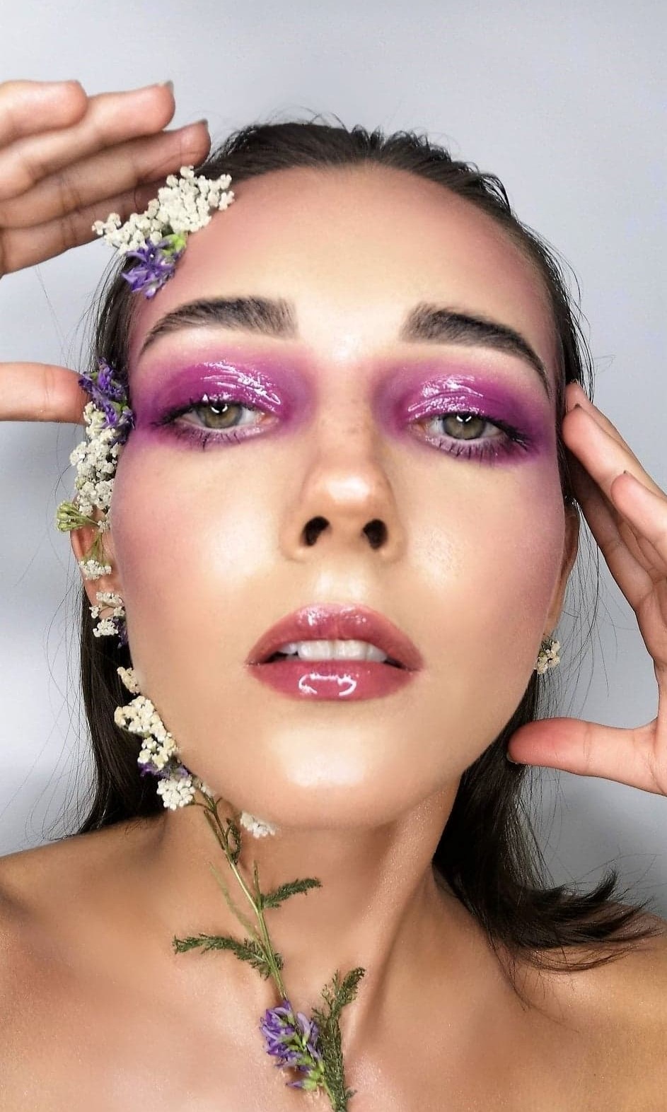 Portret kobiety z intensywnym, fioletowym makijażem oczu z efektem błyszczącego lakieru, ozdobionym drobnymi białymi i fioletowymi kwiatami, delikatny błyszczyk na ustach, dłonie uniesione przy...
