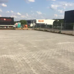 Nowo ułożona, szara kostka brukowa na placu przed budynkami firmowymi. W tle widoczny czerwony samochód dostawczy, betonowa mieszarka i przenośna toaleta.